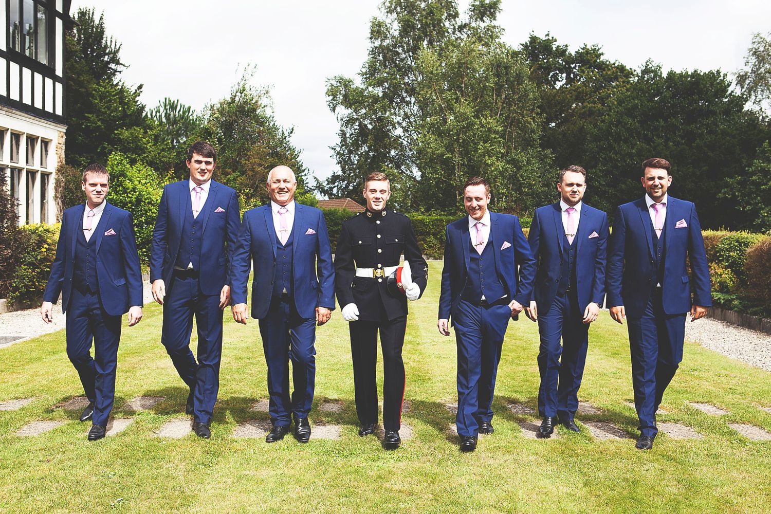 Groomsmen in navy blue suits walk together outdoors at a wedding, with one man in a black military uniform at the center.