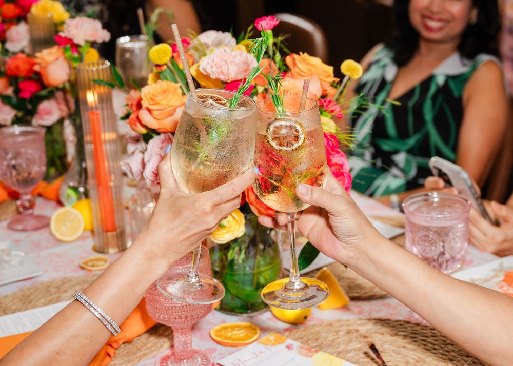 Women toasting colorful cocktails with citrus garnishes at a festive floral table setting.