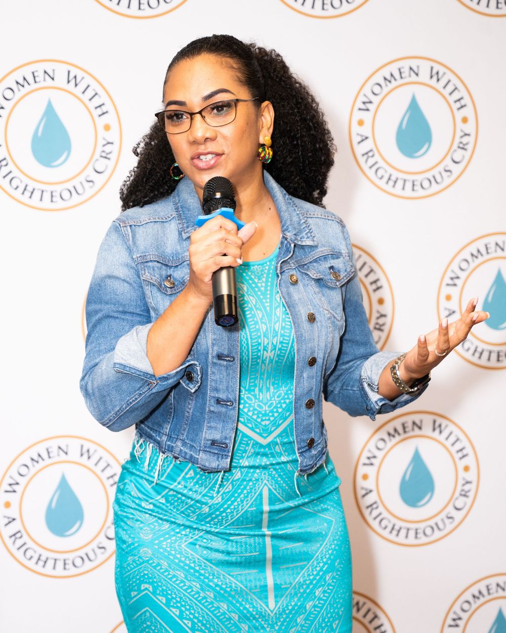 Woman in teal dress and denim jacket speaks at Women With Righteous Cry event holding microphone.