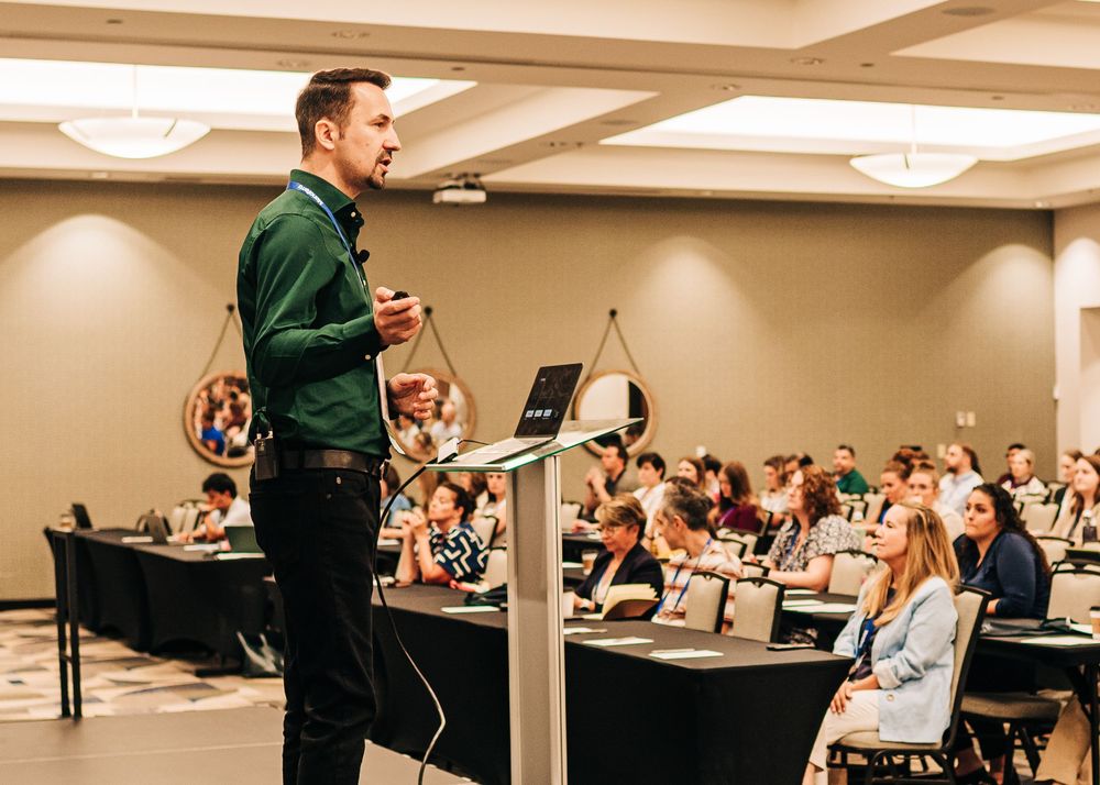 A male speaker presents at a podium to a large audience in a conference room during a professional seminar.