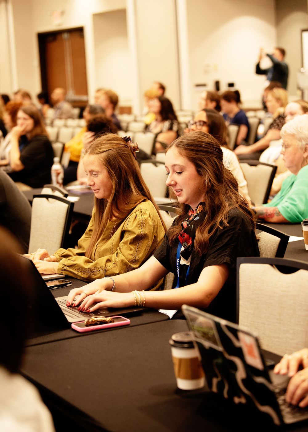 Two young women work on laptops at a conference table during a seminar with attendees seated in rows behind them.