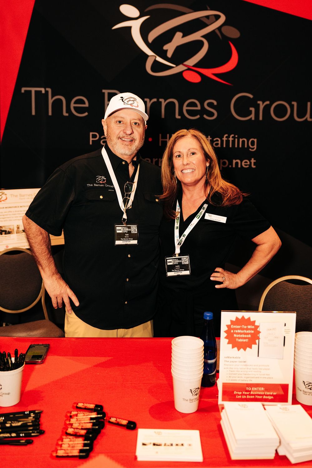 Two smiling staff members pose at The Barnes Group staffing company trade show booth with promotional materials.
