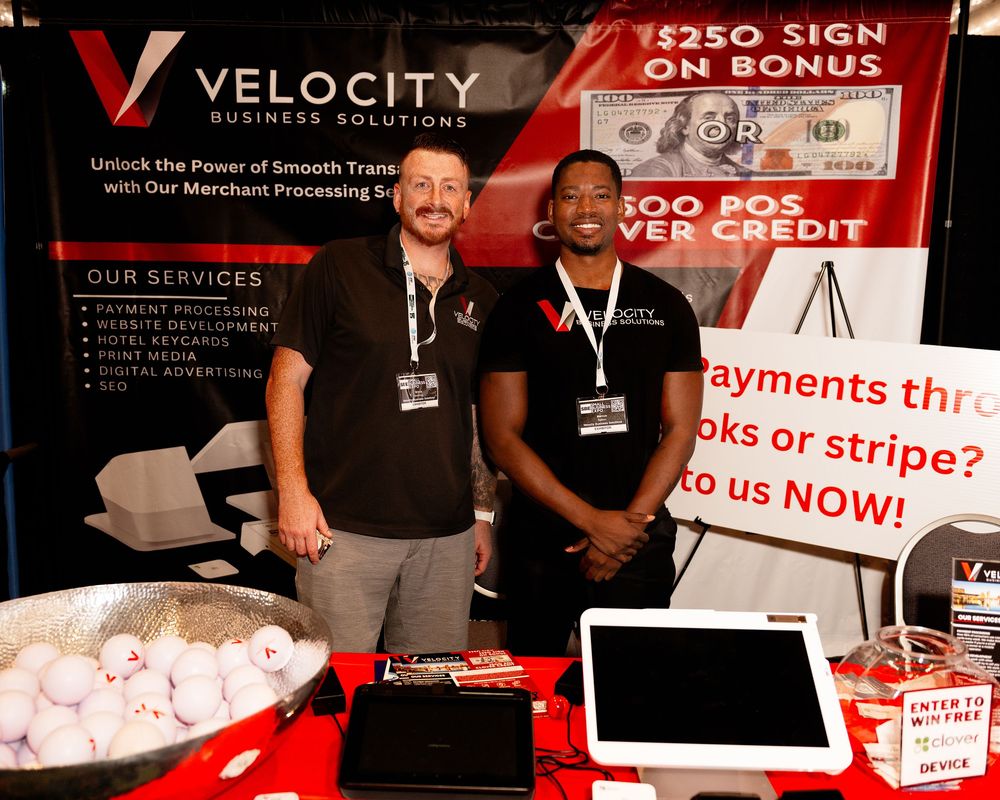 Two men smile at a Velocity Business Solutions trade show booth offering merchant processing services and a $250 sign-on bonus.