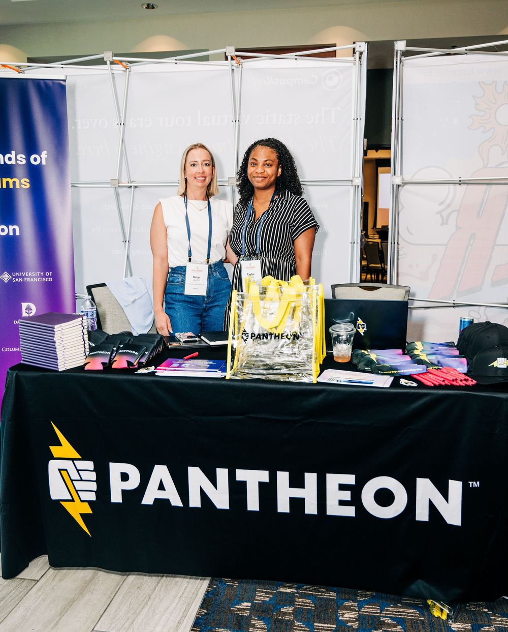 Two women smile at a Pantheon booth displaying branded merchandise and swag bags at a tech conference.