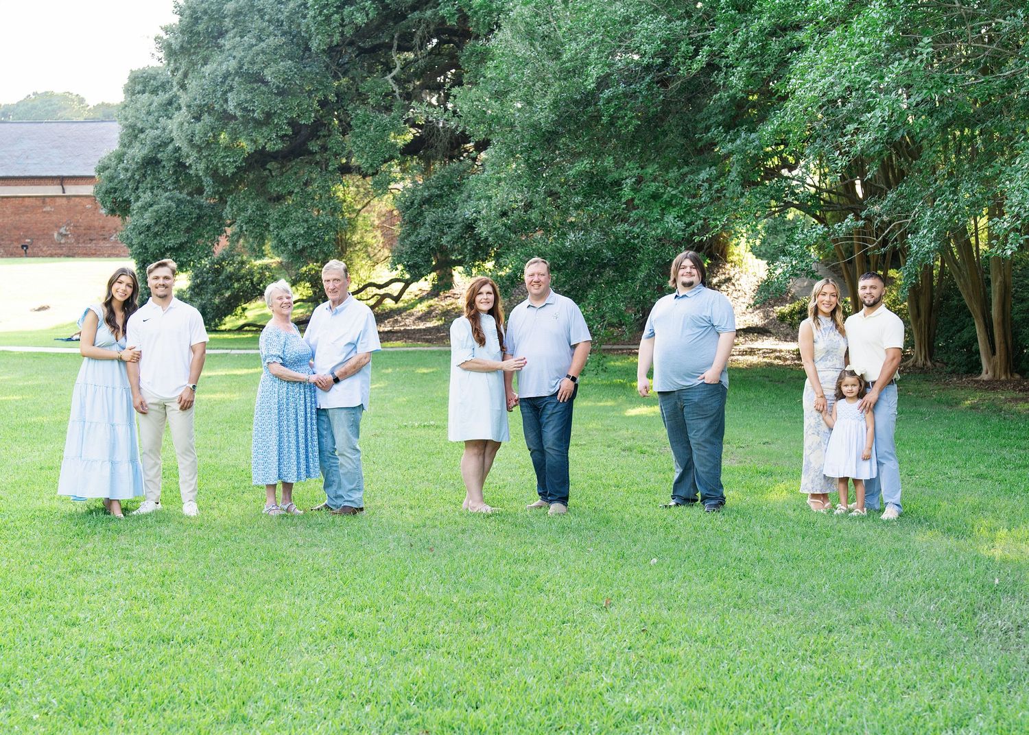 Large family group portrait on green lawn with tall trees, wearing coordinated blue and white outfits on a sunny day.