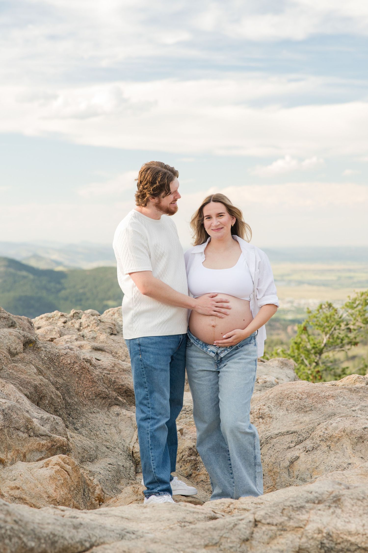 Pregnant couple standing on rocky hilltop with mountain valley views, woman in white top and jeans cradling baby bump.