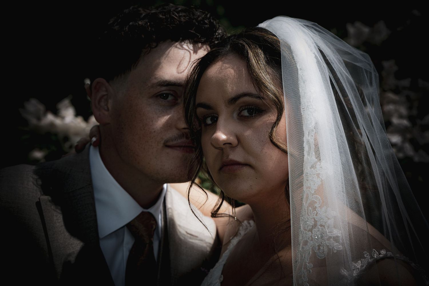 Intimate close-up portrait of bride and groom under veil surrounded by dark floral background.