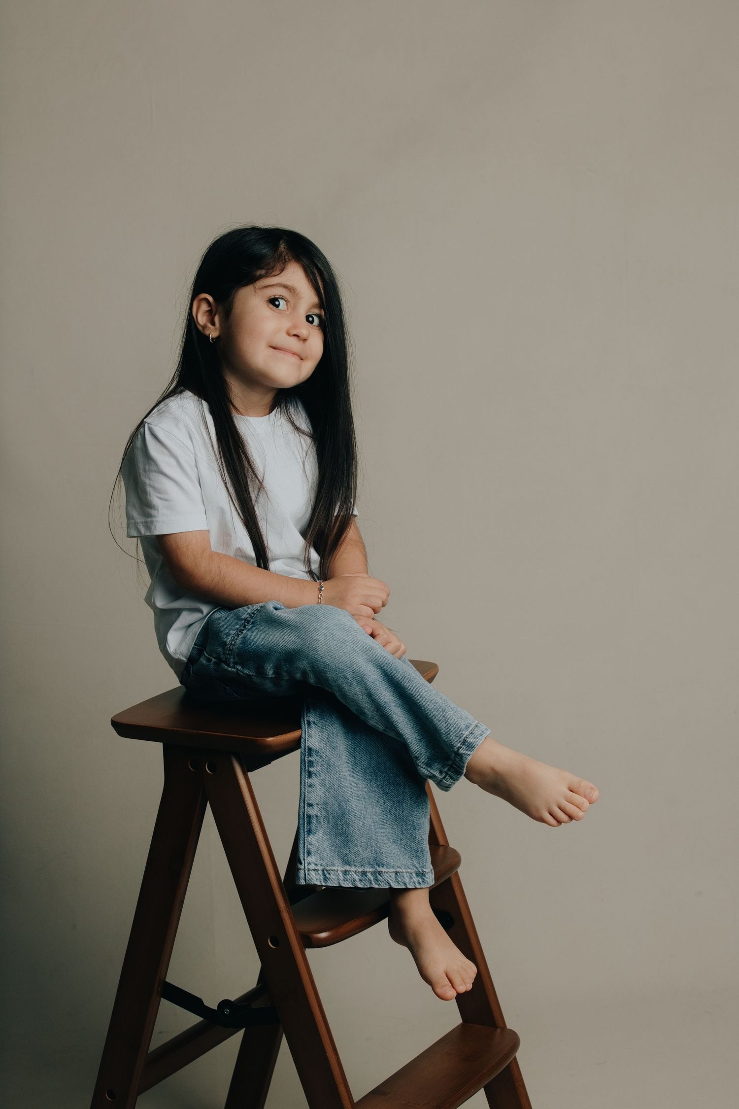 A young girl with long dark hair sits barefoot on a wooden step stool, wearing a white tee and jeans, smiling softly.