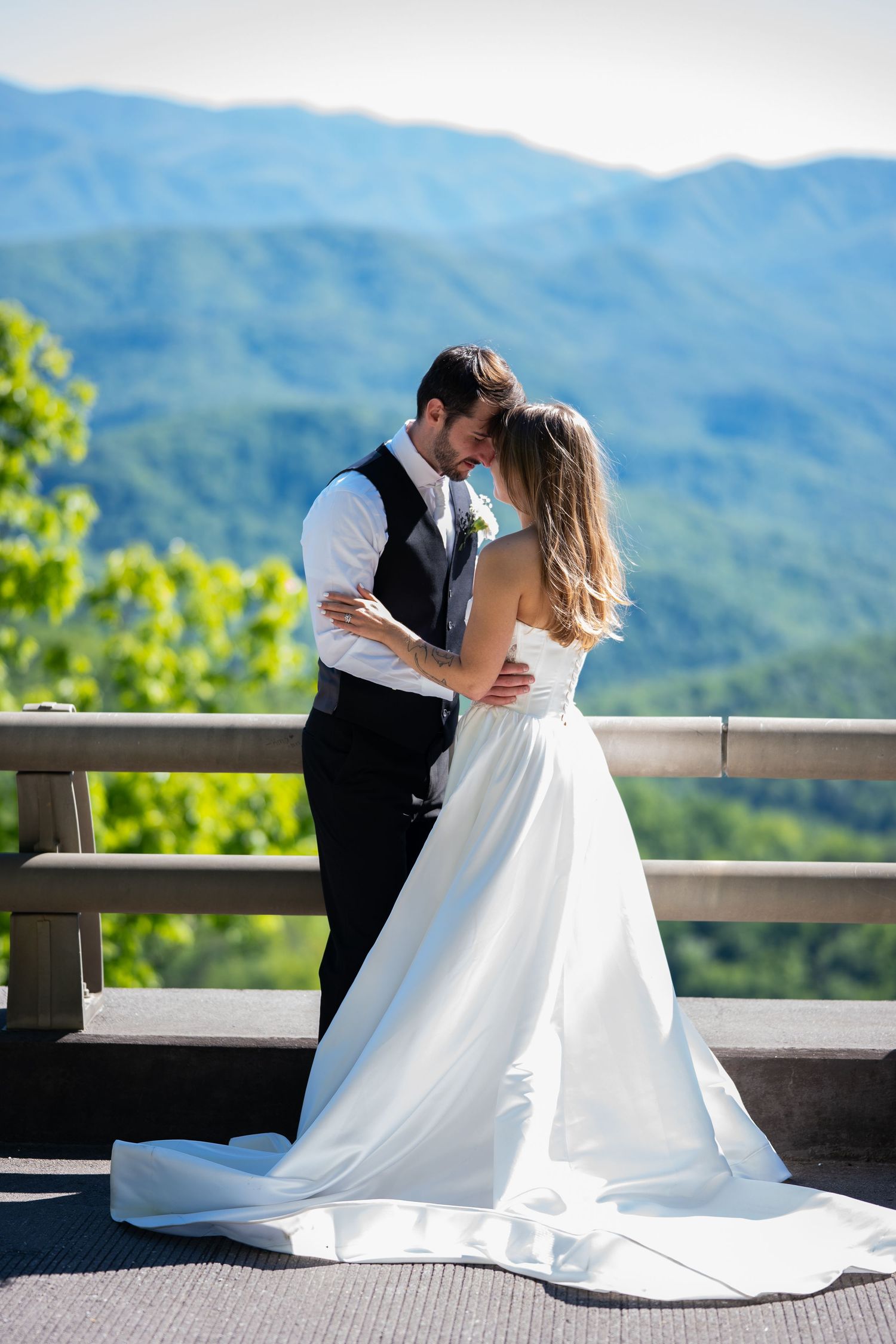 Bride and groom share a romantic moment on a mountain overlook, surrounded by lush green hills under a clear blue sky.