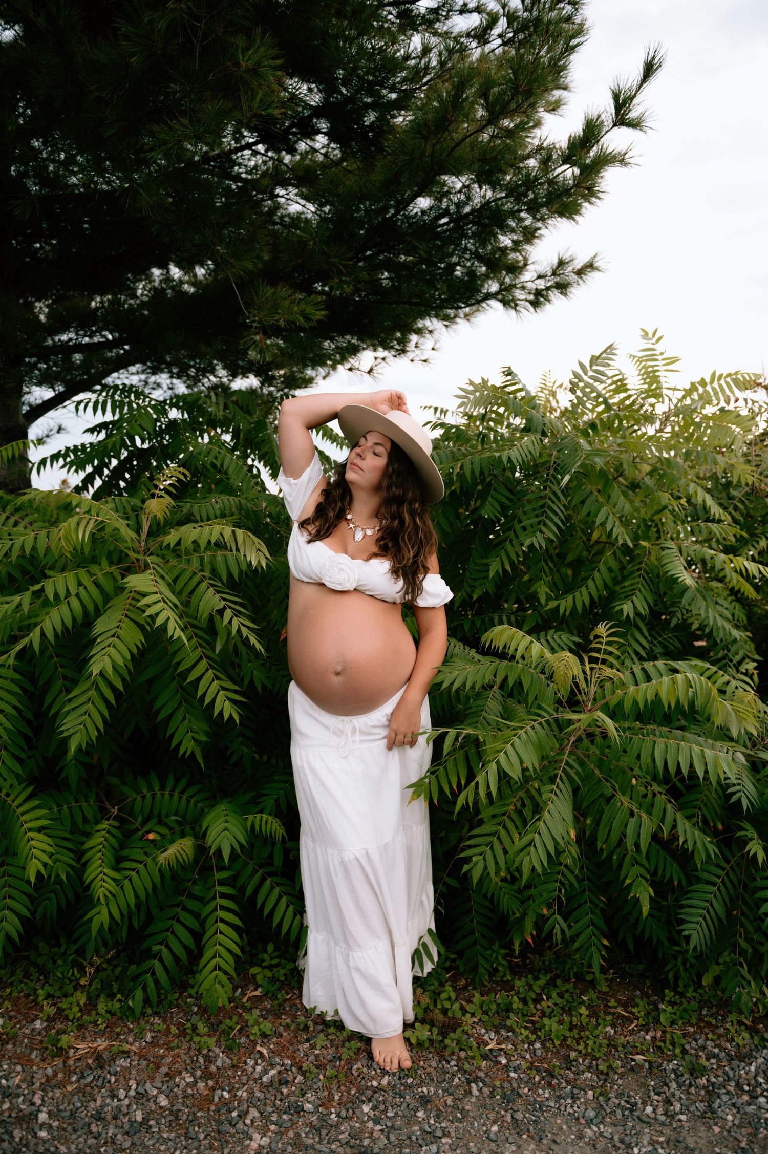 Pregnant woman in white crop top and skirt with boho hat poses barefoot among lush green ferns and pine trees outdoors.
