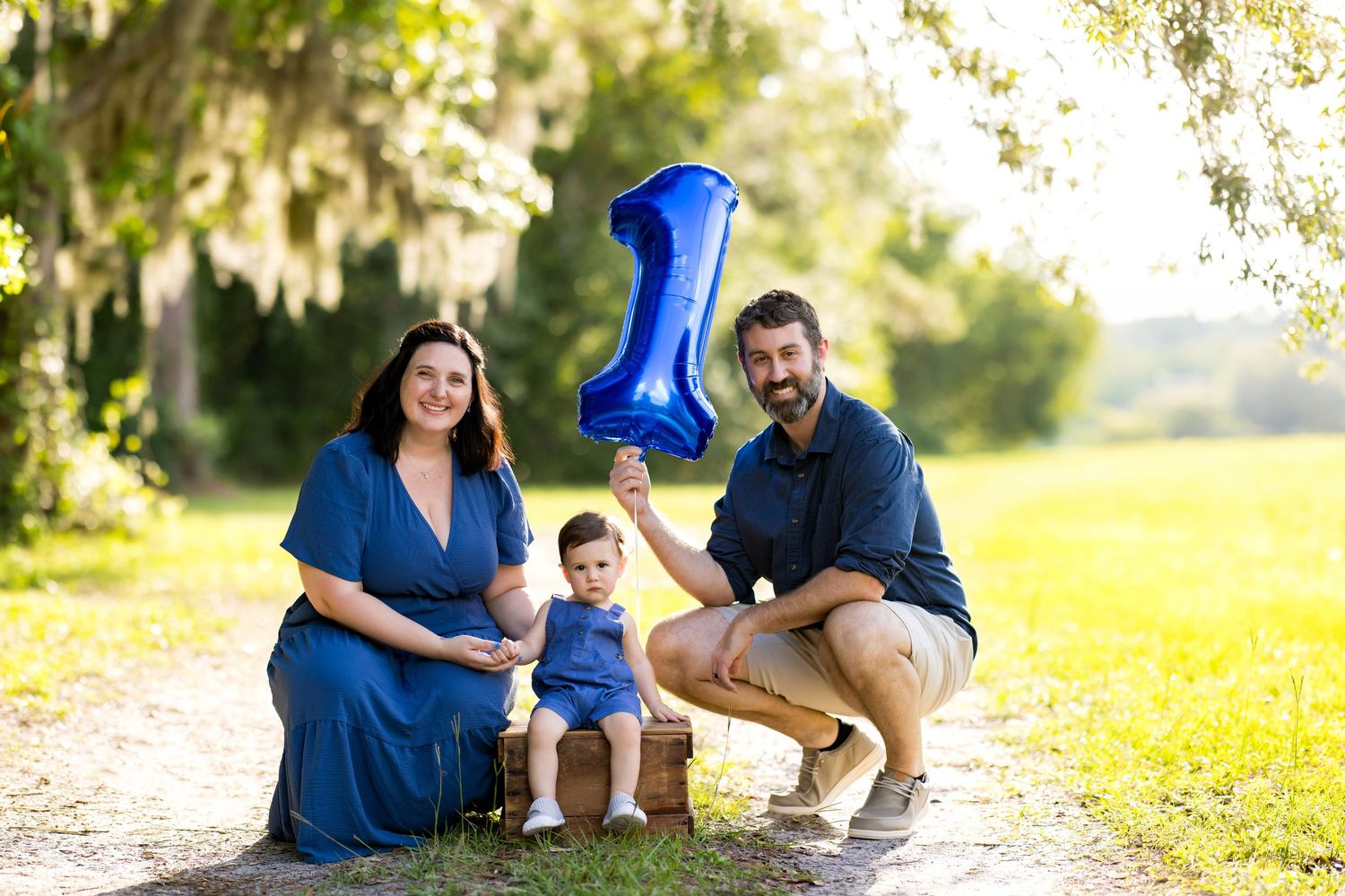 Family of three celebrating baby’s first birthday outdoors, holding a blue number one balloon in a sunny park.