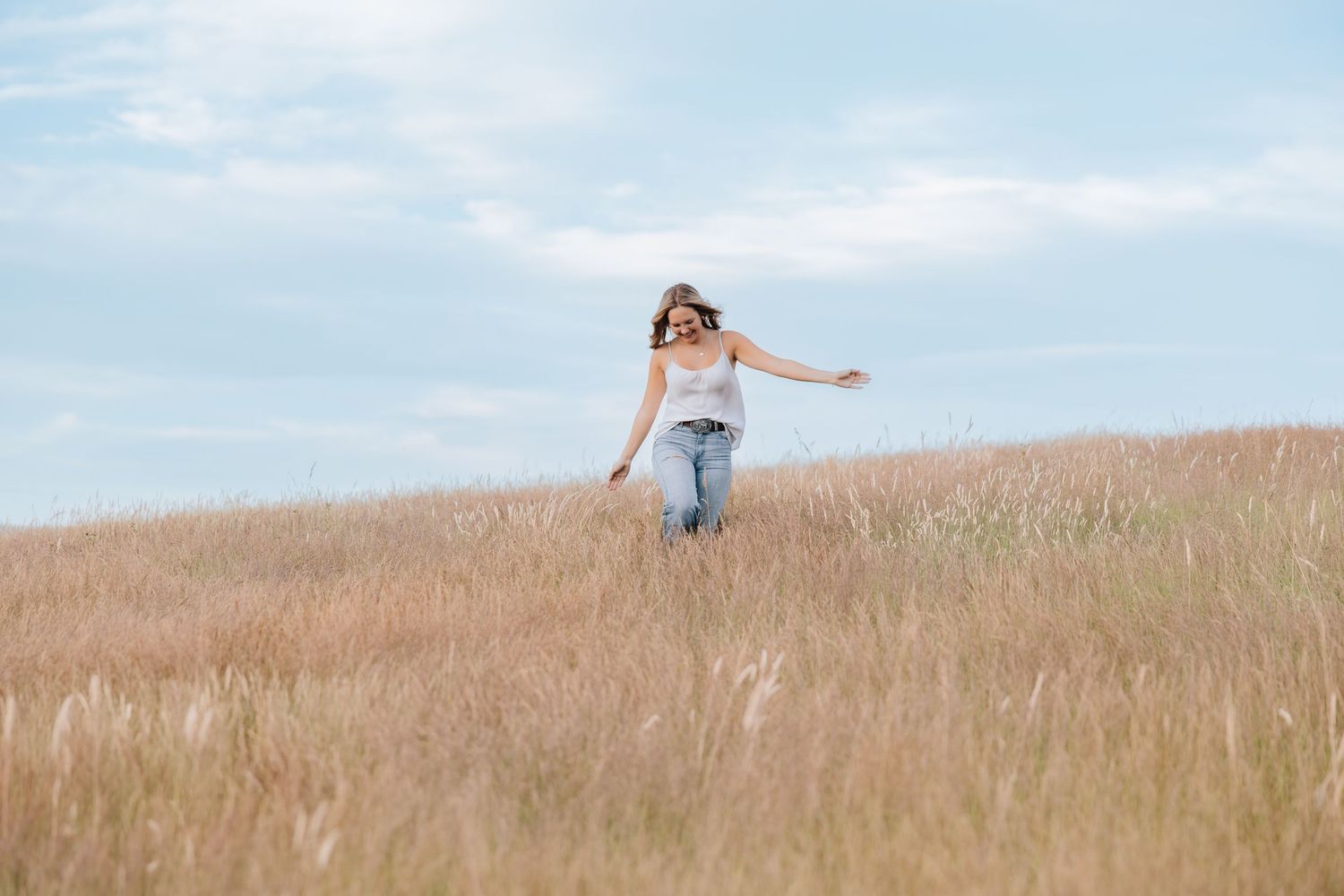 A young woman in jeans and white top walks freely through golden wheat field under blue sky.