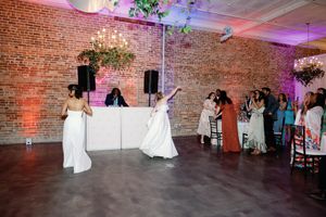 Two brides in white gowns share a first dance at a wedding reception in a brick-walled venue with warm lighting. Wilmington NC