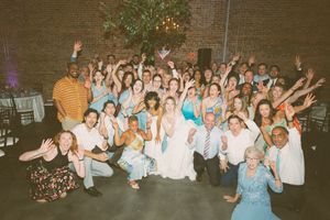 Large wedding party group photo with brides, and guests cheering with hands raised in a brick venue. LGBTQ+ Wedding Photographer