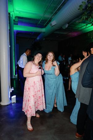 Two women in pink and blue dresses smile at a wedding reception with colorful green and purple lighting.