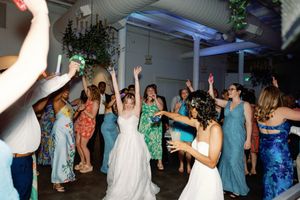 Joyful wedding reception dance floor with brides and guests celebrating with hands raised in a decorated venue. LGBTQ+ Weddings, SarahBooth