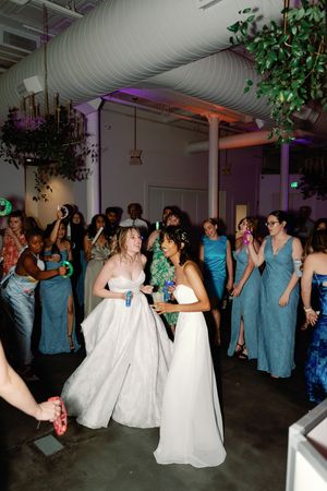 Two brides in white gowns dancing together surrounded by guests in blue dresses at a wedding reception. LGBTQ+ Weddings Wedding Photography