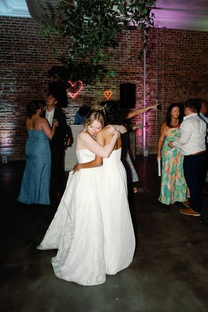 Two brides share a first dance embrace at their wedding reception with heart neon lights and guests in background. Wilmington NC Wedding