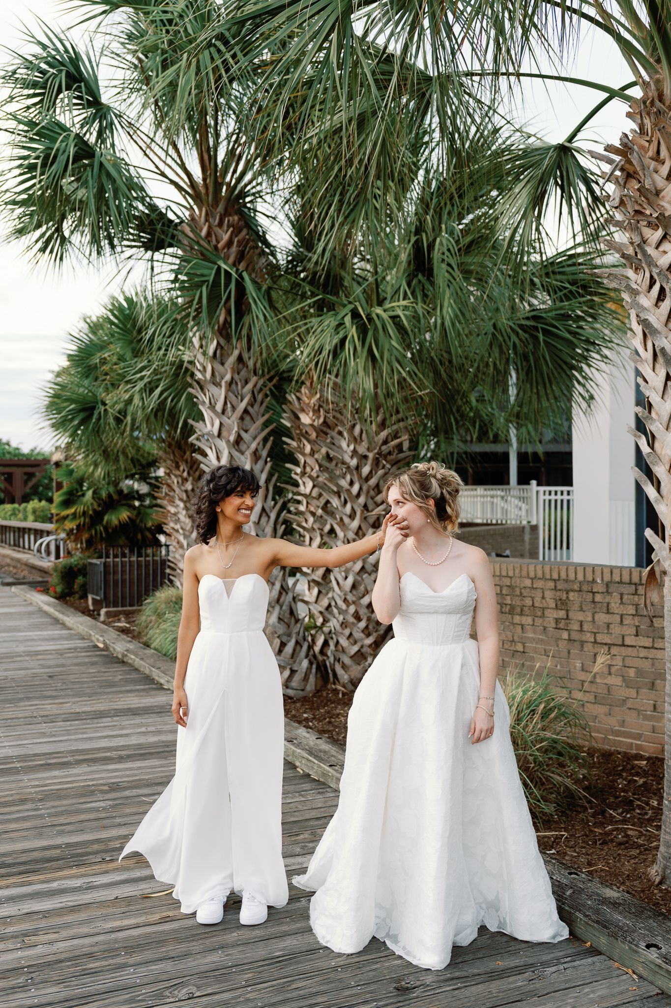 Two brides in white strapless wedding gowns walk along a boardwalk near tropical palm trees.