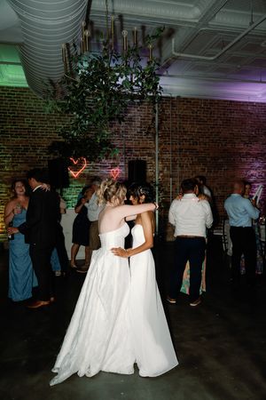 Two brides share a romantic dance at their wedding reception in a rustic brick venue with neon heart decorations. The Elia Wedding Venue