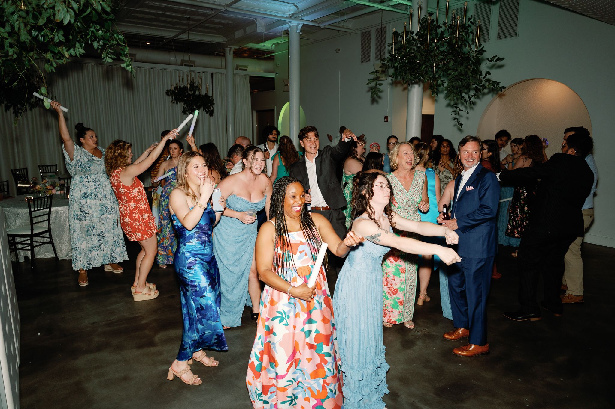 Guests dancing joyfully at an elegant wedding reception with lush greenery decor and colorful formal attire. Downtown Wilmington, NC