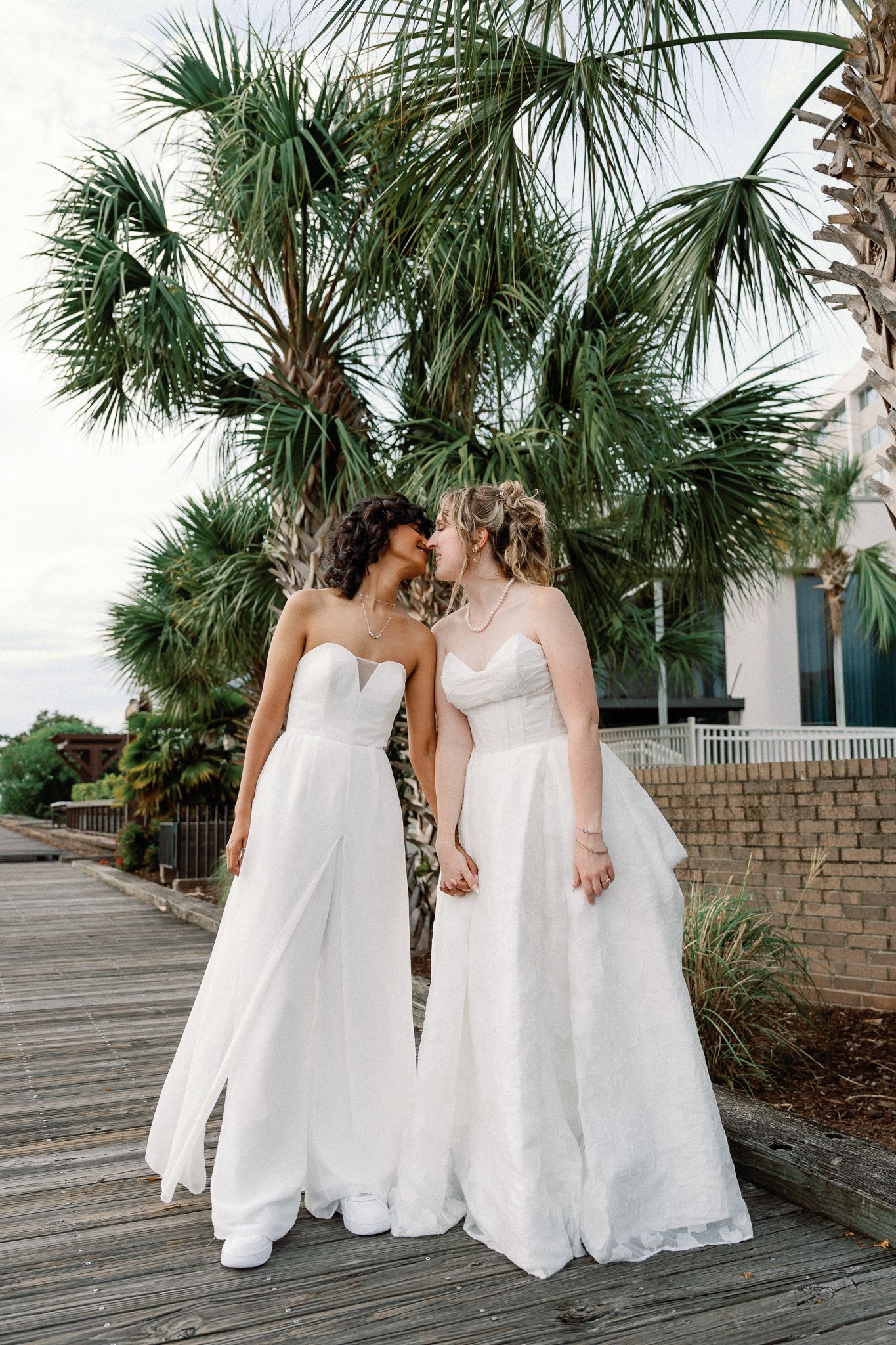 Two brides in white gowns share a kiss on a boardwalk surrounded by palm trees at a beach wedding.