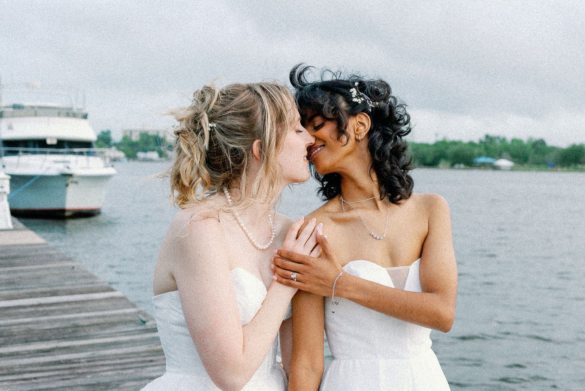 Two brides in white dresses share a romantic moment on a waterfront dock on their wedding day.