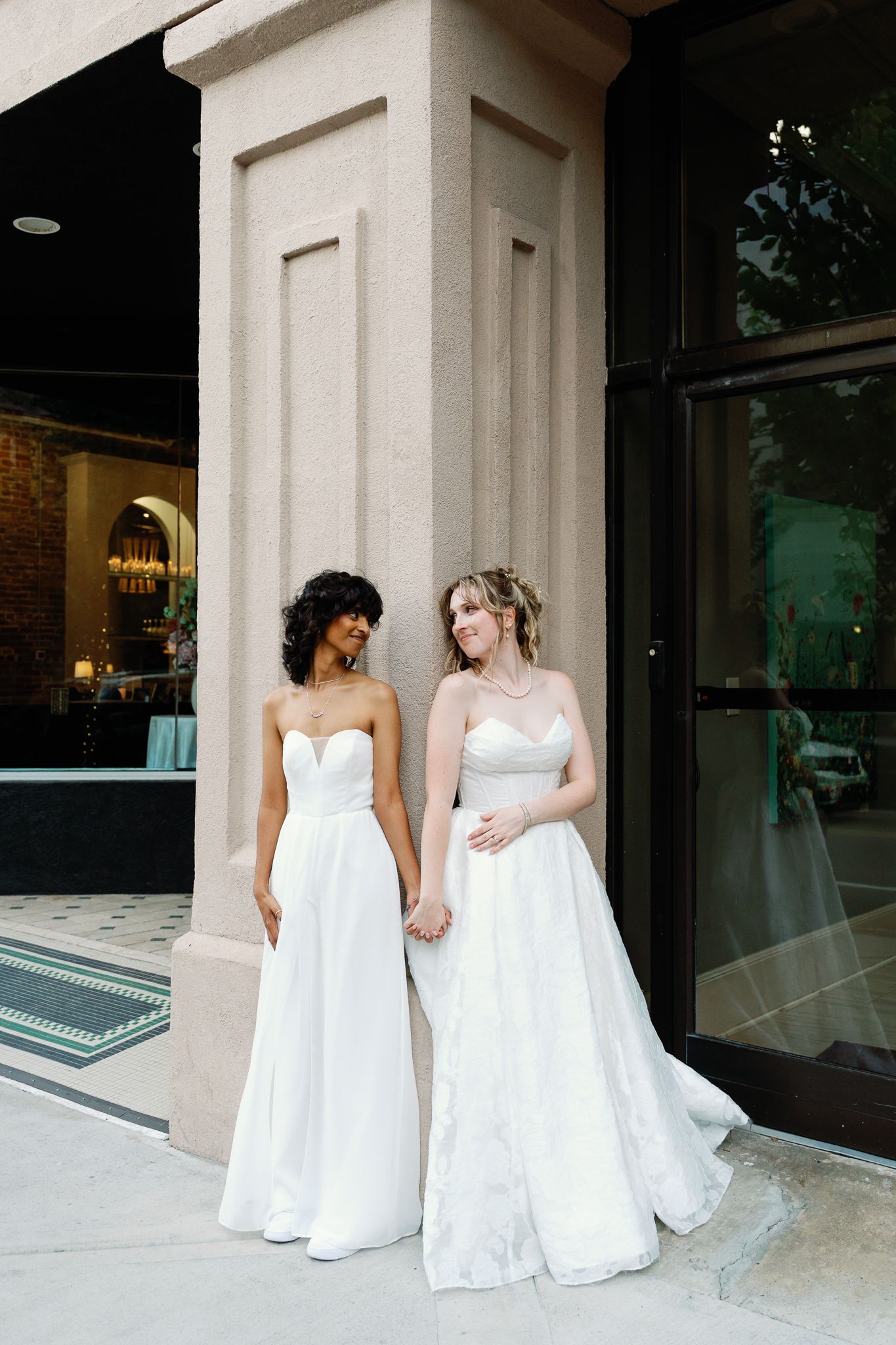 Two brides in strapless white ball gowns hold hands outside an elegant venue with stone columns.