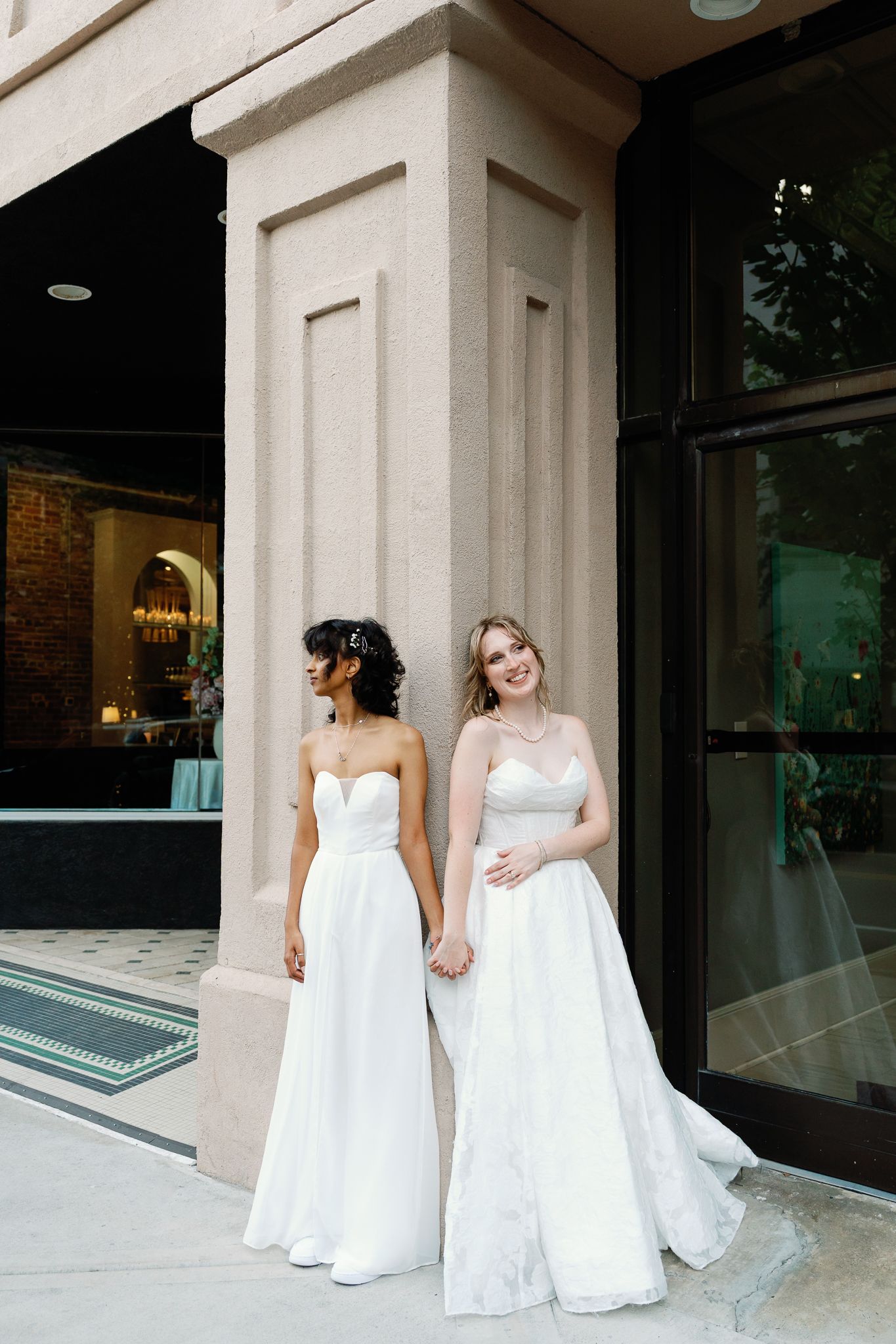 Two brides in elegant white strapless gowns stand back-to-back outside a classic stone building.