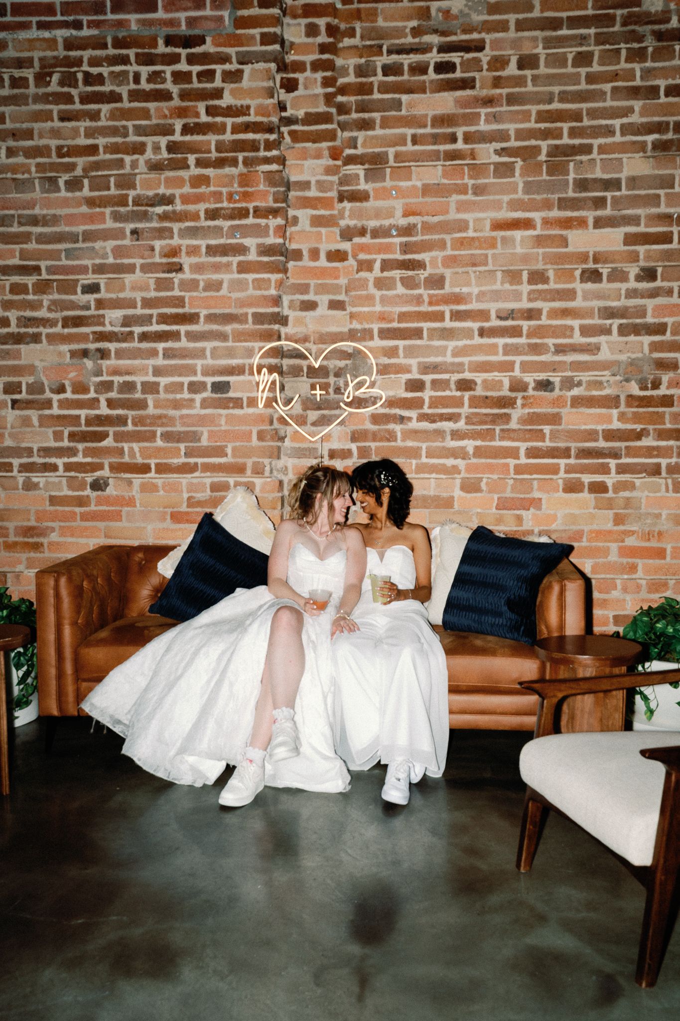 Two brides in white dresses sit closely on a leather sofa against a brick wall with a neon heart sign.