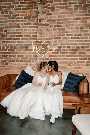 Two brides in white gowns share a kiss on a leather sofa beneath a neon heart sign against a brick wall.
