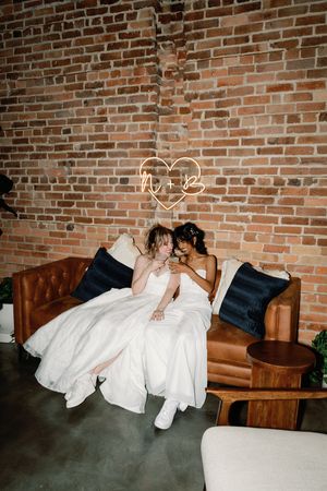 Two brides in white gowns sit closely on a leather sofa beneath a neon heart sign against a brick wall.