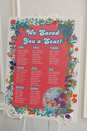 Colorful floral wedding seating chart sign reading We Saved You a Seat with numbered table assignments on pink background.