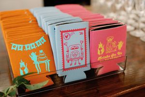 Colorful wedding koozies in orange, blue, and pink displayed in a holder, featuring fun illustrated designs as party favors.