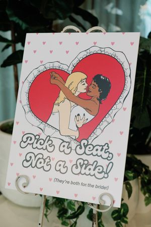 Wedding seating sign reading Pick a Seat Not a Side featuring retro illustrated couple embracing in a heart.