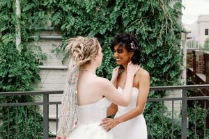 Two brides in white strapless gowns share a joyful moment on a vine-covered balcony.