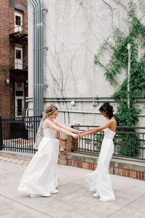 Two brides in white gowns hold hands and dance together on an urban rooftop terrace on their wedding day.