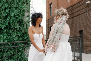 Two brides in white gowns laugh together outdoors, one wearing a floral ribbon veil.