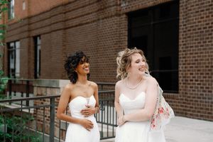 Two brides in white strapless gowns laughing together outdoors near a brick building on their wedding day.