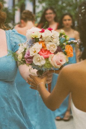 Bridesmaids in blue dresses surround a colorful bridal bouquet of roses and mixed flowers at a wedding.