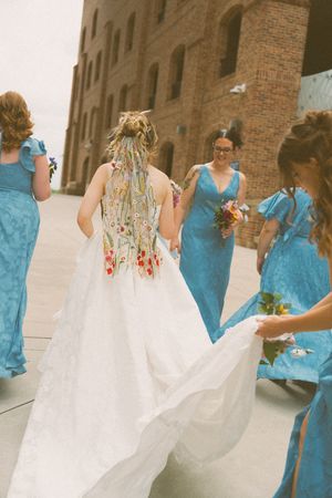 Bride in embroidered white gown walks with bridesmaids in blue dresses, train held, near brick building.