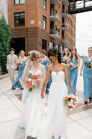 Two brides in white gowns hold colorful bouquets, walking with wedding party in blue dresses outside brick building.