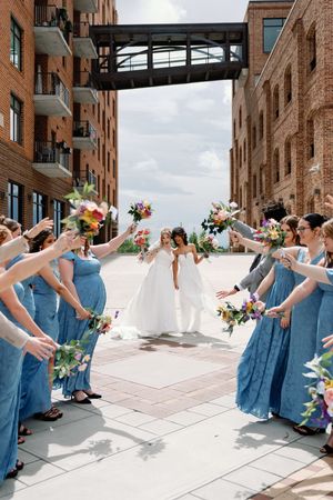Two brides walk between bridesmaids in blue dresses holding colorful bouquets aloft at an outdoor urban wedding venue.