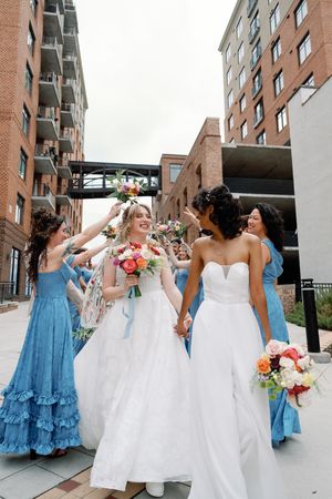 Two brides in white gowns walk with bridesmaids in blue dresses, holding colorful bouquets outside a brick building.