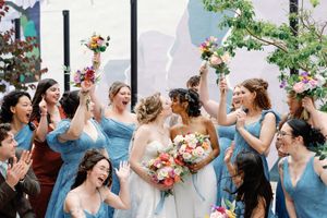 Two brides share a kiss surrounded by cheering bridesmaids in blue dresses holding colorful bouquets at a joyful wedding.