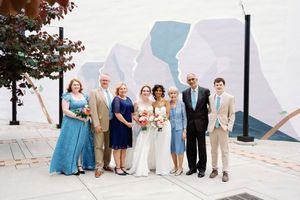 Wedding party poses before a colorful mural outdoors, featuring two brides in white and guests in blue and tan attire.