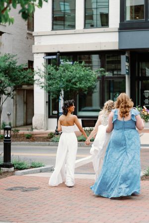 Three women in wedding attire walk hand-in-hand across a brick crosswalk in an urban setting.