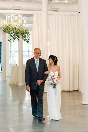 Bride in strapless white gown walks with her father down the aisle in an elegant white venue with chandeliers.