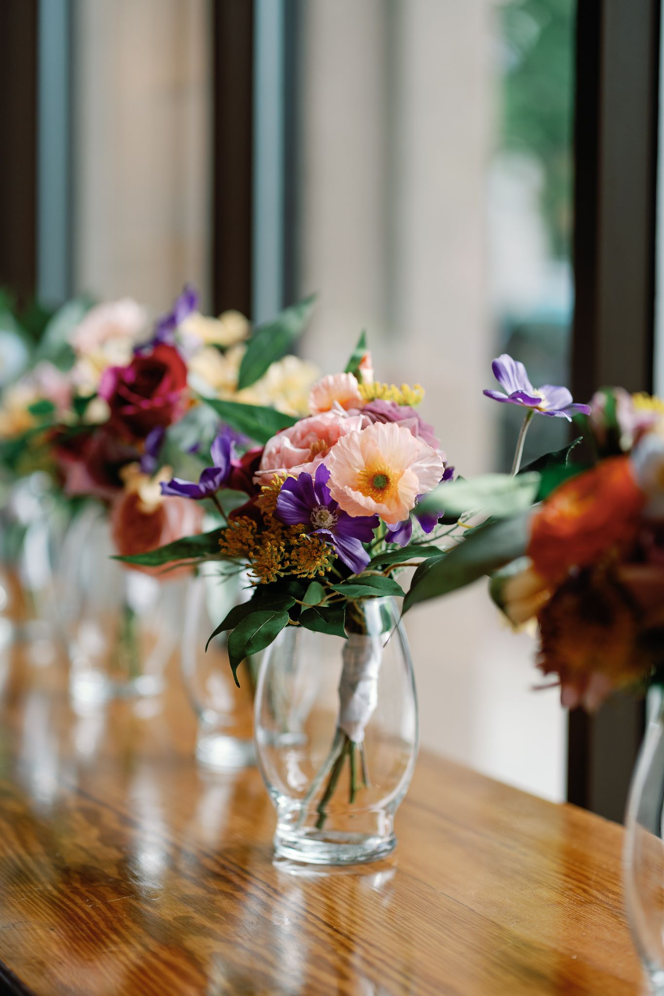 Colorful floral arrangements in clear glass vases lined up on a wooden table near a window.