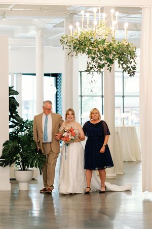 Bride walks down aisle with parents in elegant white venue with chandelier and greenery at wedding ceremony.